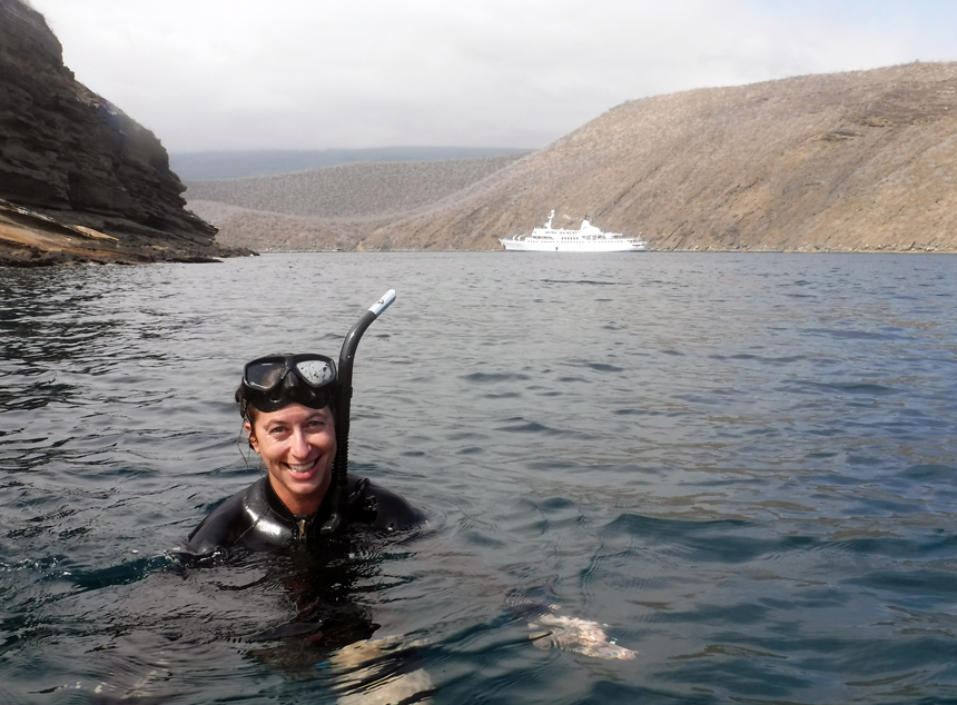 Woman in black wetsuit, snorkel & goggles smiles with head above water with small white ship in the distance in Galapagos.