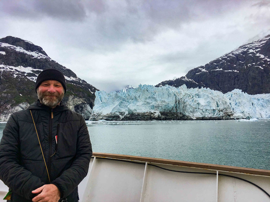 A man in a black jacket and hat standing on the deck of a ship with a tidewater glacier and mountains in the background with a cloudy sky.