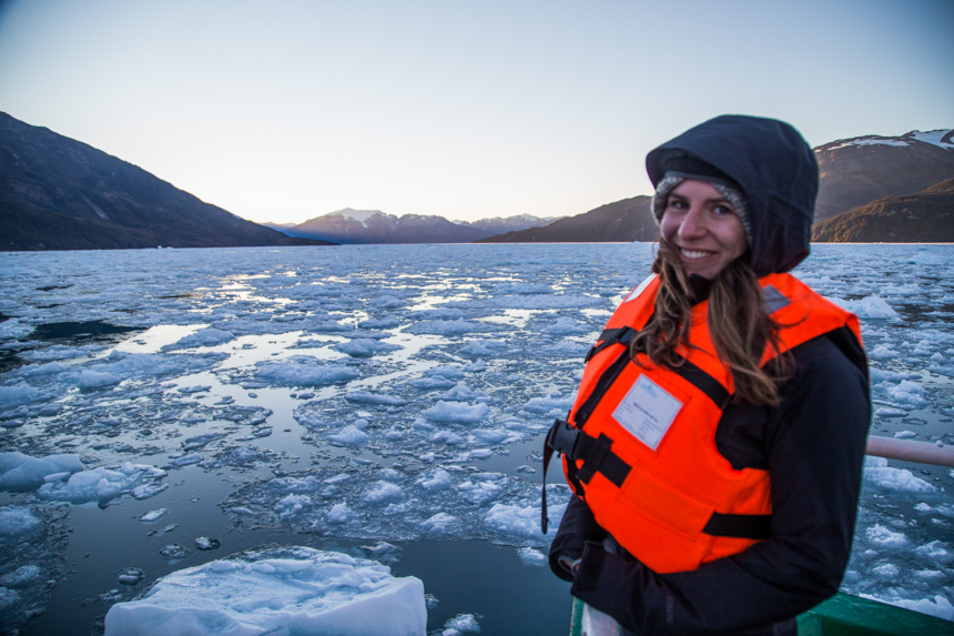 Woman in orange life jacket & dark gray coat stands among floating iceberg bits at dusk on a Patagonia small ship cruise.