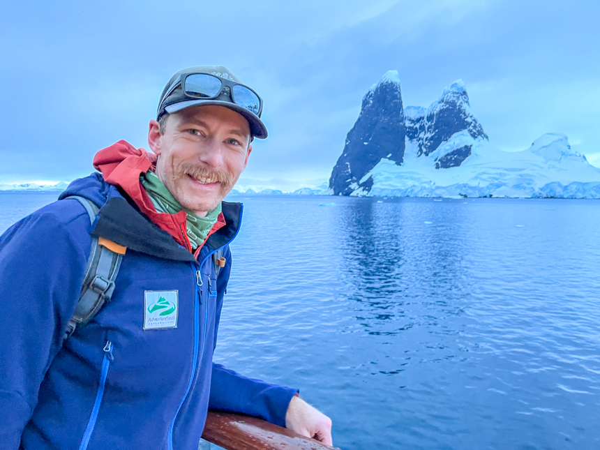 A man in a blue jacket stands near the railing on a ship's deck with an icy mountain behind him