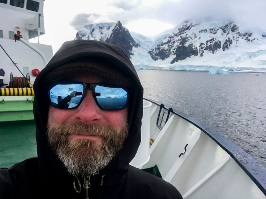 Man in sunglasses & black jacket stands on deck of Ortelius expedition ship sailing by snowy peaks in Antarctica.
