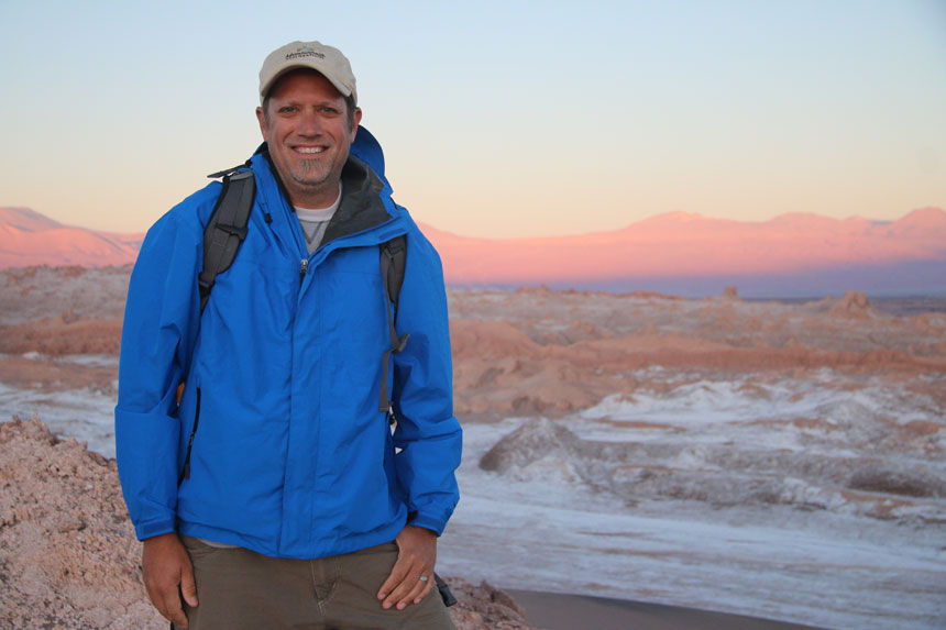Man in blue jacket & tan hat stands atop a peak in the desert at sunset with pink & purple-hue mountains behind.