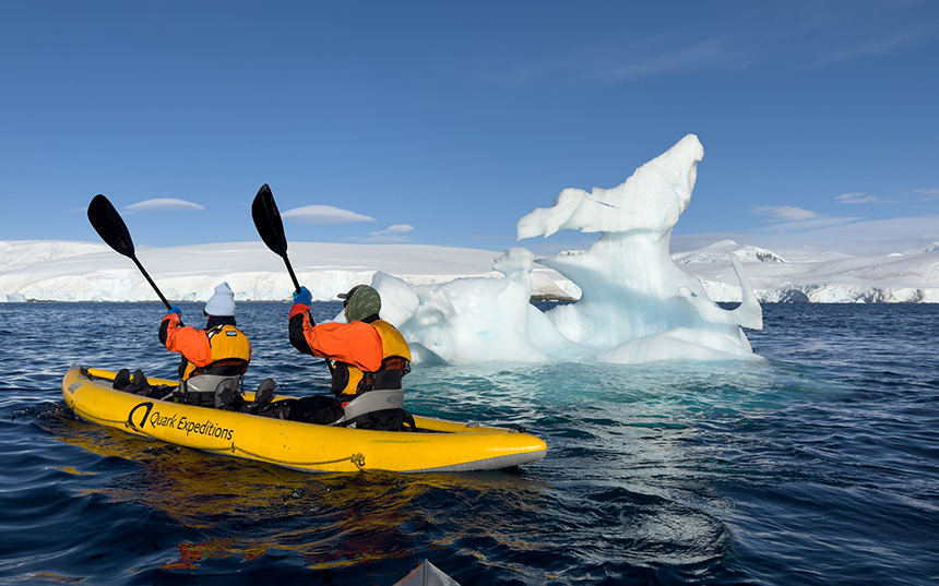 Two travelers in a yellow kayak paddle near a large iceberg in Antarctica