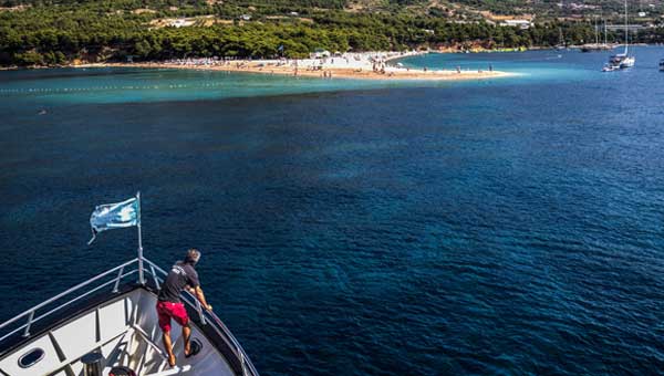 A man in red shorts stands on the bow of a ship with a blue flag heading toward a white sand beach