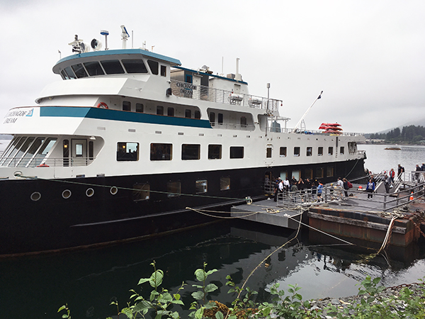 A small ship with a black hull seen tied up next to a dock on a cloudy day with passengers loading on the ship.