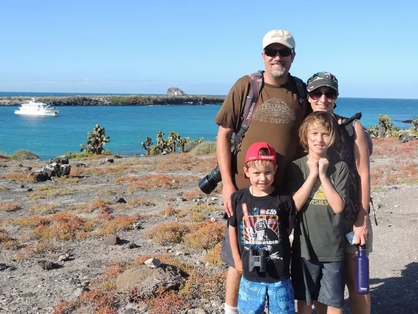 Husband & wife stand with 2 boys on sandy island with red bushes & green cacti with small ship behind in Galapagos.