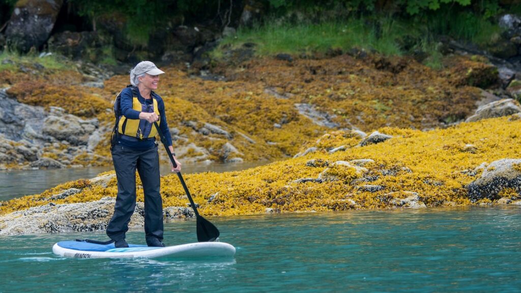 a single female traveler seen on a stand-up paddleboard in Alaska with bright moss on the shoreline behind her