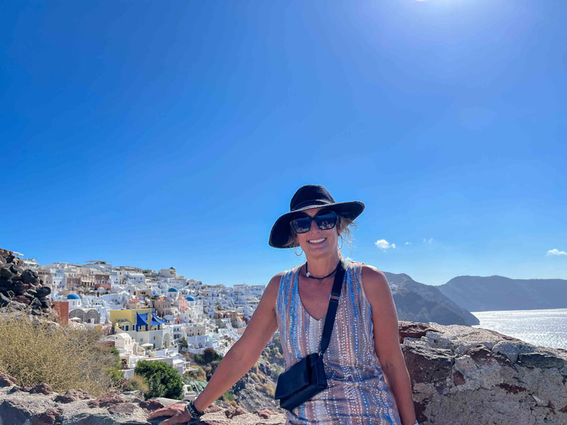 A female traveler in a hat and sunglasses standing in front of a Greek island village with many white houses looking over the Mediterranean Sea.