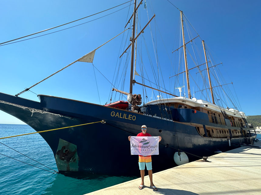 Man in red t-shirt & yellow & teal shorts holds a white flag with the AdventureSmith Explorations logo in front of Galileo blue-hulled motorsailor.