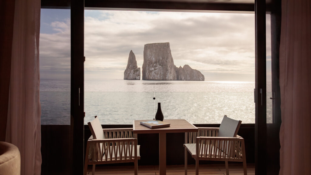 Suite private balcony at dusk on Hermes mega catamaran with table, 2 padded chairs & glass railing overlooking Kicker Rock.