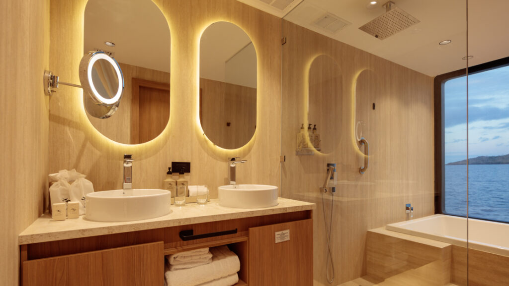 Guest bathroom with stone tub by window, glass-walled rain shower, vanity & dual bowl sinks on Hermes Galapagos catamaran.