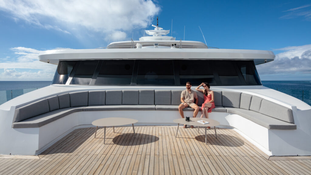 Man & woman sit in sun on gray padded bench of forward-facing teak decking on Hermes Galapagos luxury catamaran.
