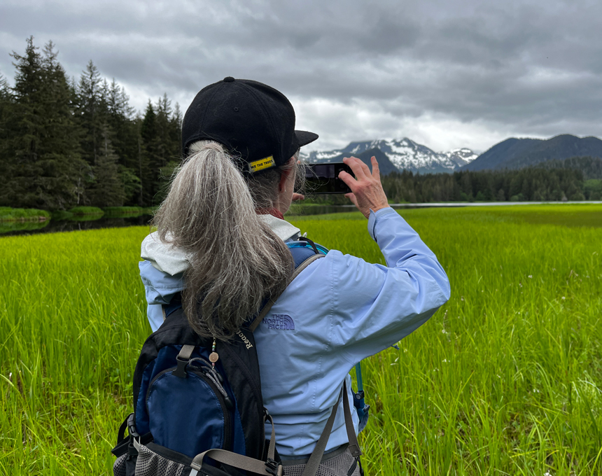 A woman with gray hair and a black baseball cap, blue jacket and backpack seen from behind taking an iphone photo of green grass and snowcapped peaks in Alaska