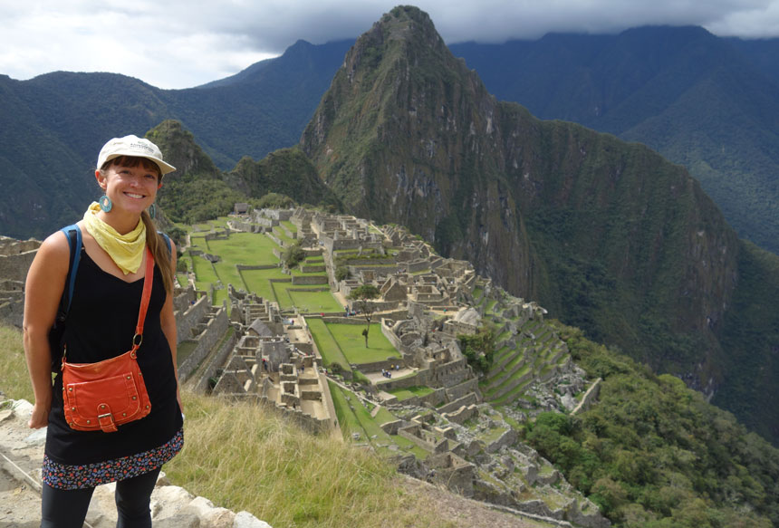 Woman in black dress, tan hat, yellow scarf & red handbag stands overlooking Machu Picchu with green terraced grass & ancient stone walls.