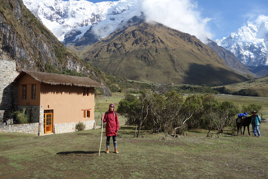 Woman in red jacket & knitted hat holds a walking stick while standing in front of an adobe & stone building in the high mountains.