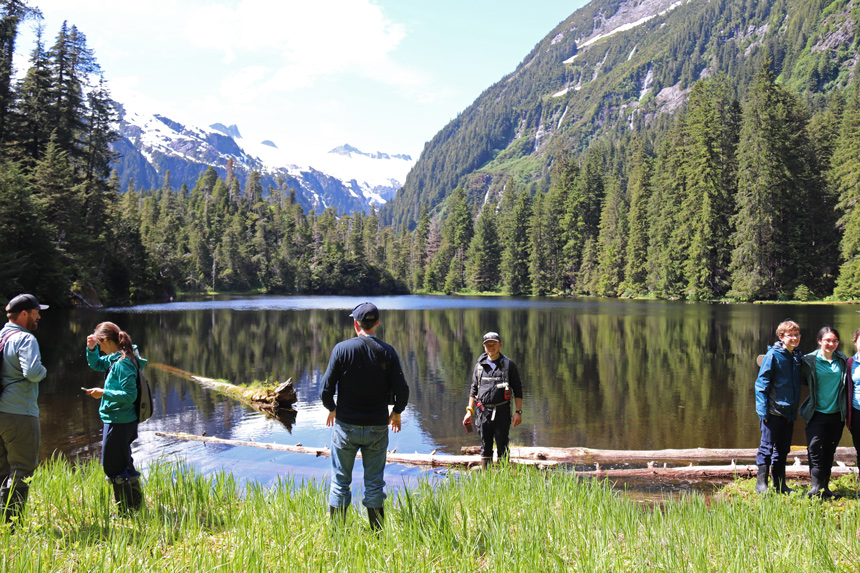 At the end of a bushwhacking activity UnCruise guests stand and take photos in front of an alpine lake surrounded by trees and mountain range.