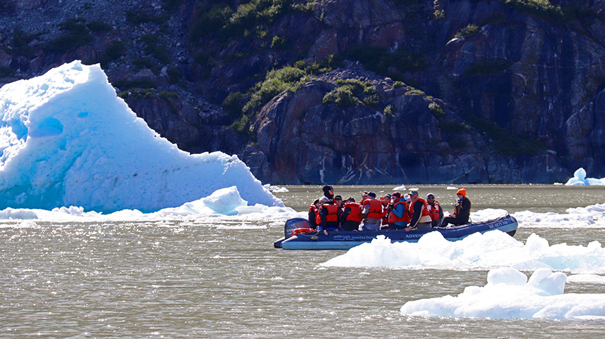An inflatable skiff filled with guests in orange life jackets cruises between floating icebergs in Alaska.