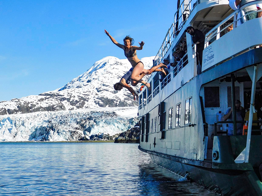 Two cruise guests take polar plunge, jumping to the water from their ship in front of an icy glacier and snowy mountain range in Alaska.