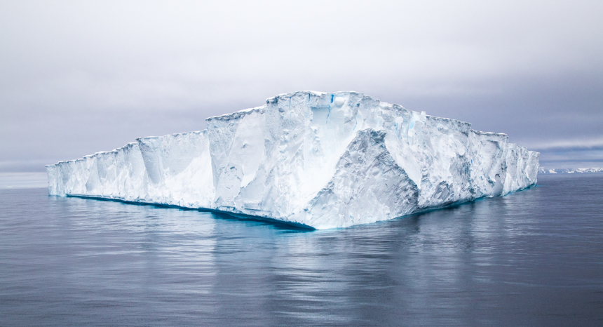 A massive white flat topped tabular iceberg the size of a house floats in the dark blue ocean of Antarctica in November.
