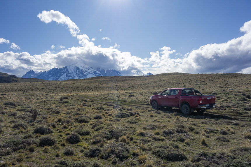 a red truck seen in a field with snowcapped mountains topped with clouds