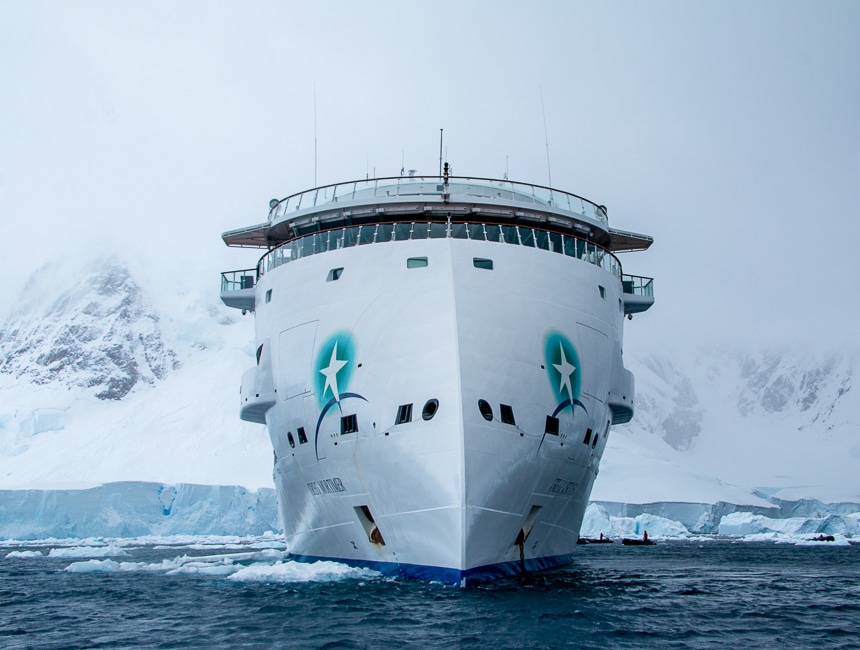 An all white futuristic and modern ship cruises the dark ocean water in front of a mist covered snowy mountain range in Antarctica.