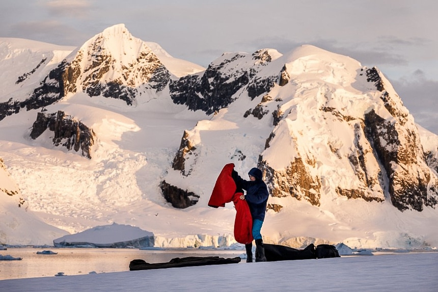 In front of a snow covered mountains cape a female guests sets up her sleeping pad and and bag as part of a polar camping activity in Antarctica.