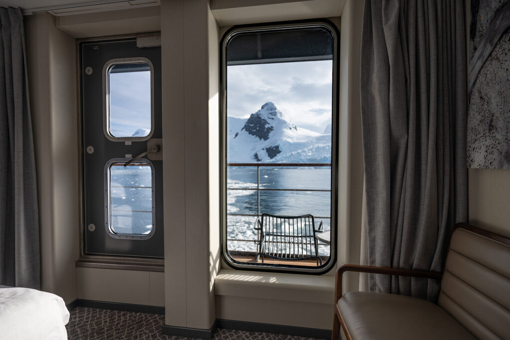 Veranda Cabin aboard Magellan Explorer showing large window and door to the private balcony. Snow covered jagged mountains show outside.