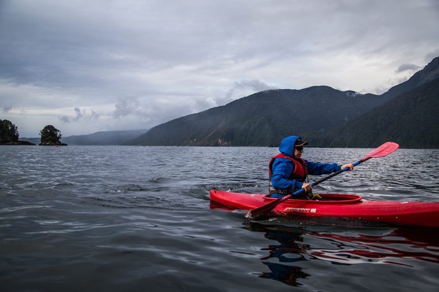 Man in rain gear & life jacket paddles a red kayak in calm sea beside dense green mountains under cloudy skies in New Zealand.