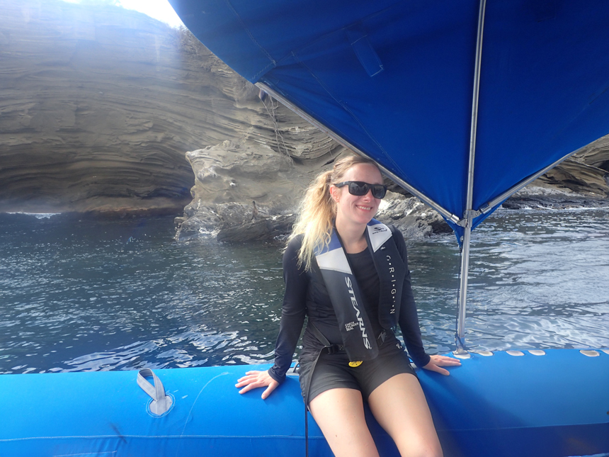 Woman in black sunglasses & shirt sits under shade on blue Zodiac boat with beige volcanic cliffs behind on a Galapagos cruise.