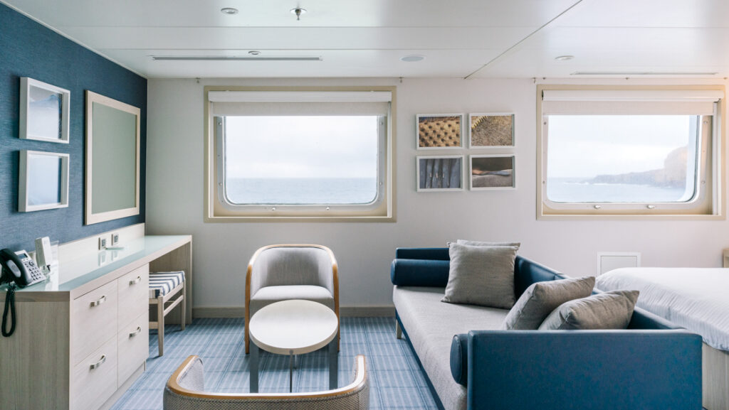 Living room area of Darwin Suite on Santa Cruz II Galapagos ship with chaise style couch, 2 chairs, wood coffee table & dresser.