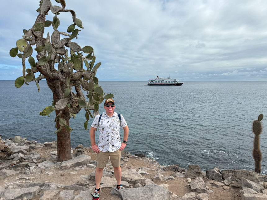 Man in white shirt & beige shorts stands beside cactus-like tree at sea shore with white, red & black ship behind in Galapagos.