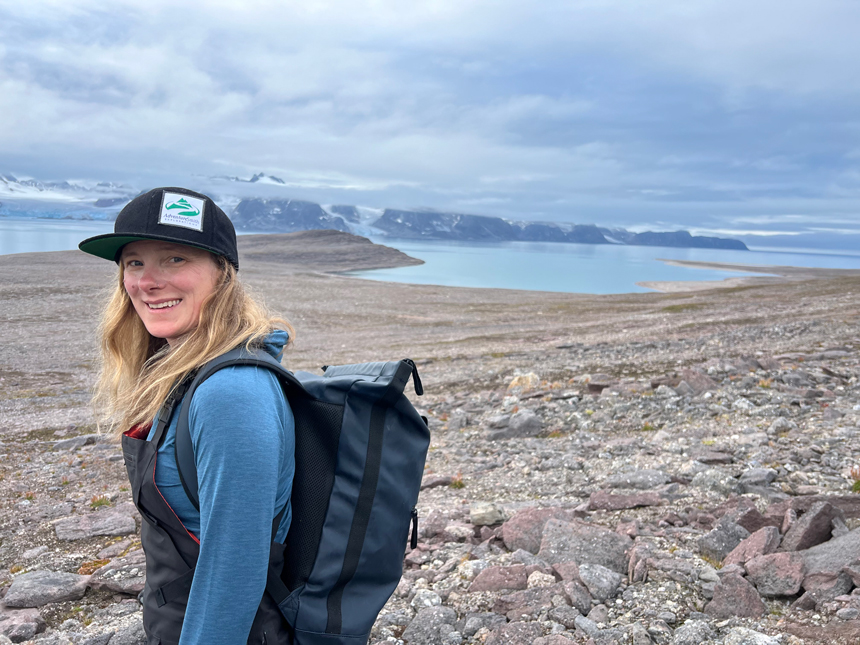 Blonde woman in black hat & blue shirt hiking over rocky ground with snowy peaks & teal sea behind in the Arctic.