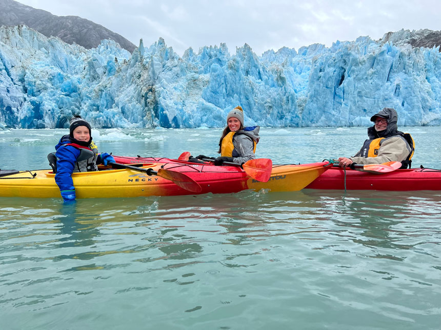 Young family paddles 2 red & yellow kayaks in front of an icy glue glacier on an Alaska Uncharted cruise.
