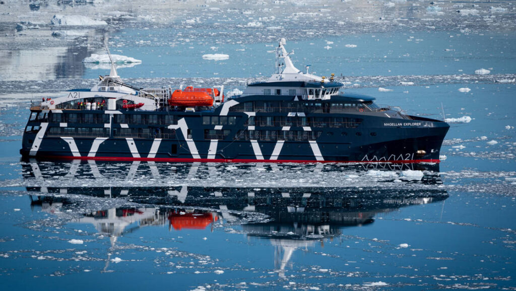 Exterior starboard side of dark navy blue Magellan Explorer Antarctica expedition ship floating on icy reflective ocean line.