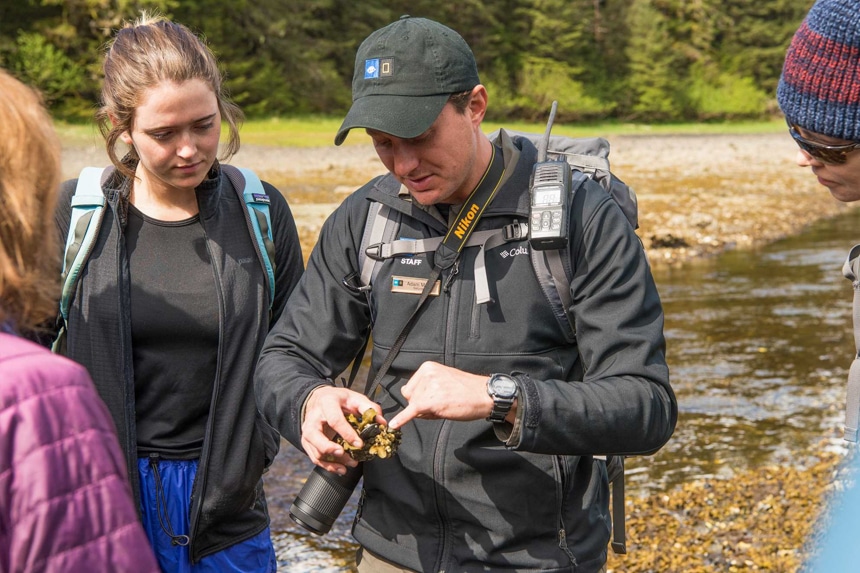 A Lindblad Expeditions naturalist guide is surrounded by cruise guests as he discusses an item he holds in his hands.