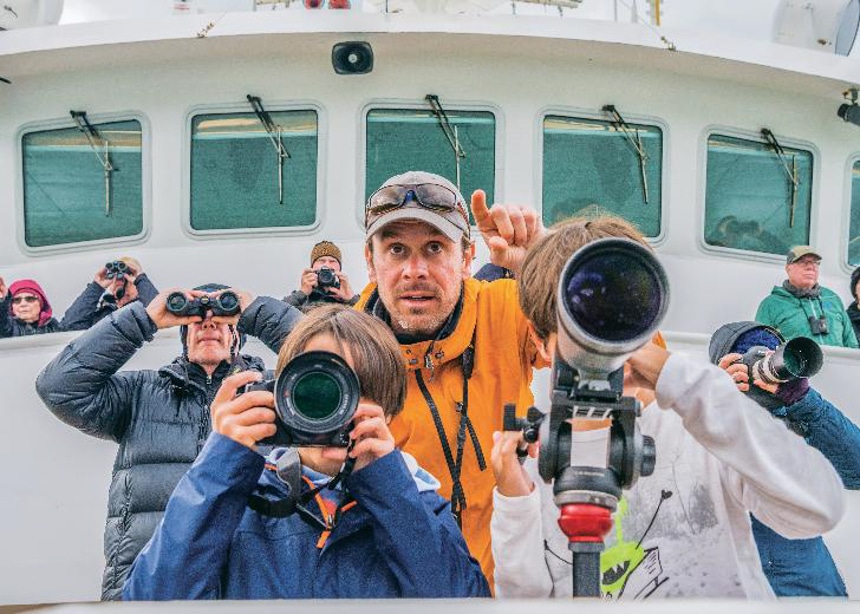 At the bow of a boat, children use cameras and binoculars as they are instructed by a Lindblad Expeditions professional photographer.