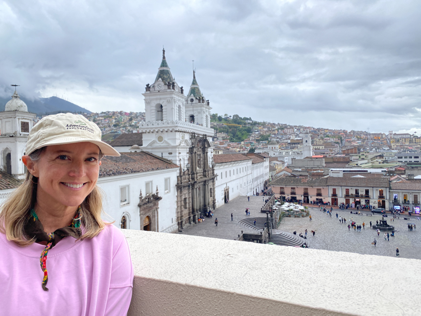 Lis Larson in an AdventureSmith Explorations hat posing with San Francisco Plaza in Quito, Ecuador from a balcony in Casa Gangotena Hotel