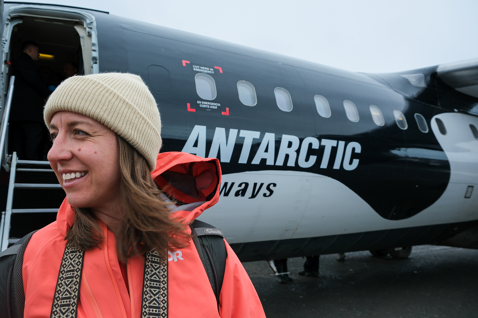 Expert in Antarctica, wearing neon pink jacket and tan beanie standing in front off Antarctic airways airplane pained in dark blue and white.