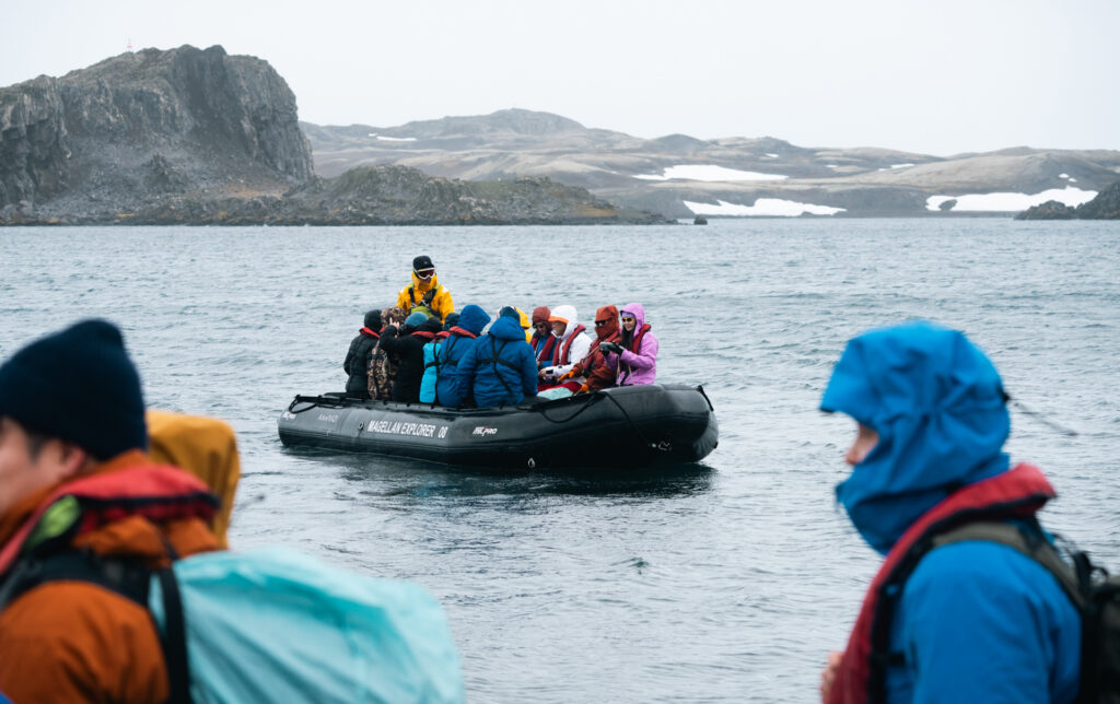 Cold, windy overcast day in Antarctica, a black inflatable skiff filled with travelers in colorful parkas sit as they navigate the ocean.