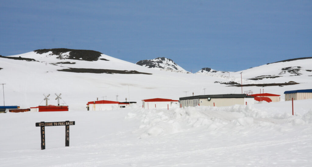 Chile's Frei Base, covered in a layer of snow, wooden sign, white buildings with red and blue roofs surrounded by melting mountainscape.