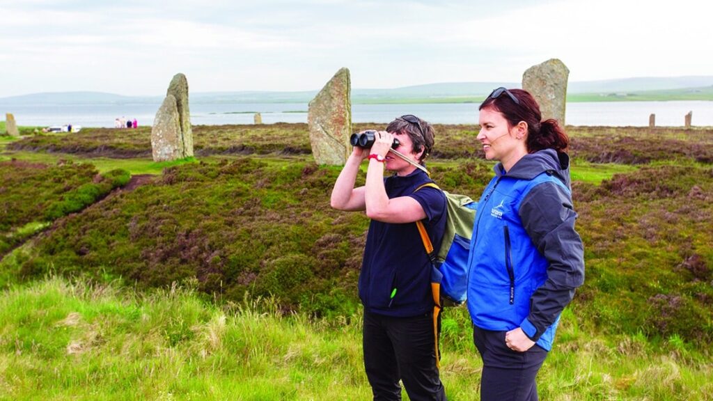 Two female travelers, one holding up binoculars to her eyes, standi n front of stone pillars at the Ring of Brodgar in Orkney, Scotland.