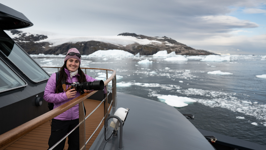 AdventureSmith Grace pictured aboard Magellan Explorer with her large camera in hand pictured with large icebergs off in the distance
