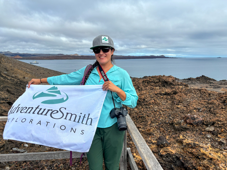 Woman in teal shirt & gray baseball cap holds white flag with green AdventureSmith Explorations logo on wood platform backed by beige volcanic rock in Galapagos.