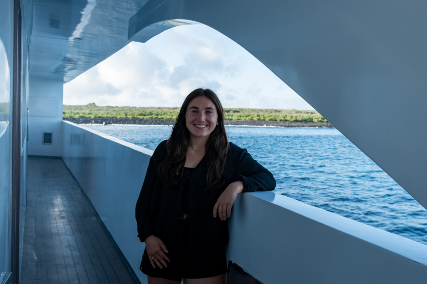 Woman in black shirt stands with arm along side of modern small ship backed by green shore in Galapagos.