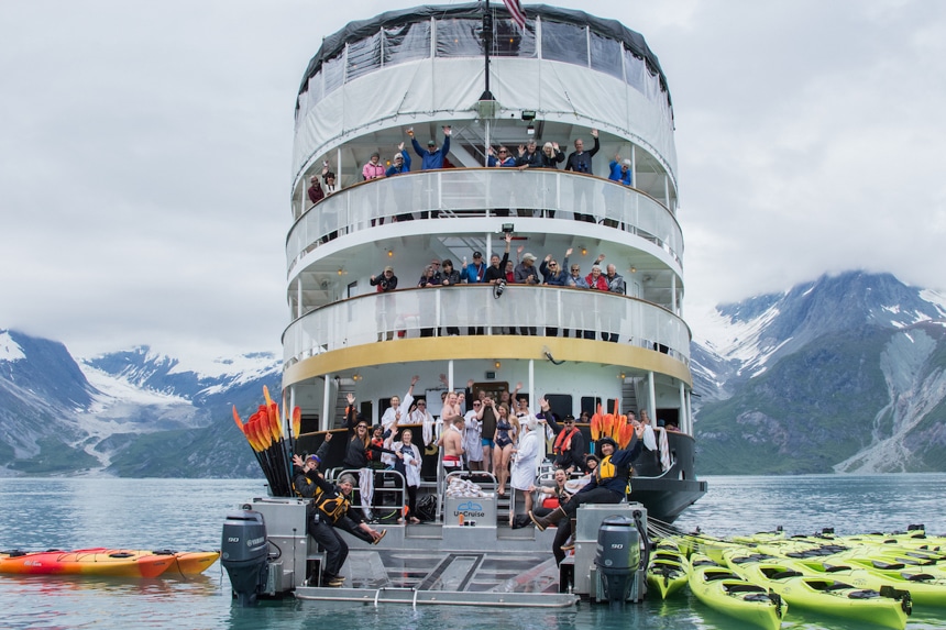 Guests and crew wave from the back of the 4 level boat, the Wilderness Adventurer, as it floats in Alaska surrounded by a mountain range.