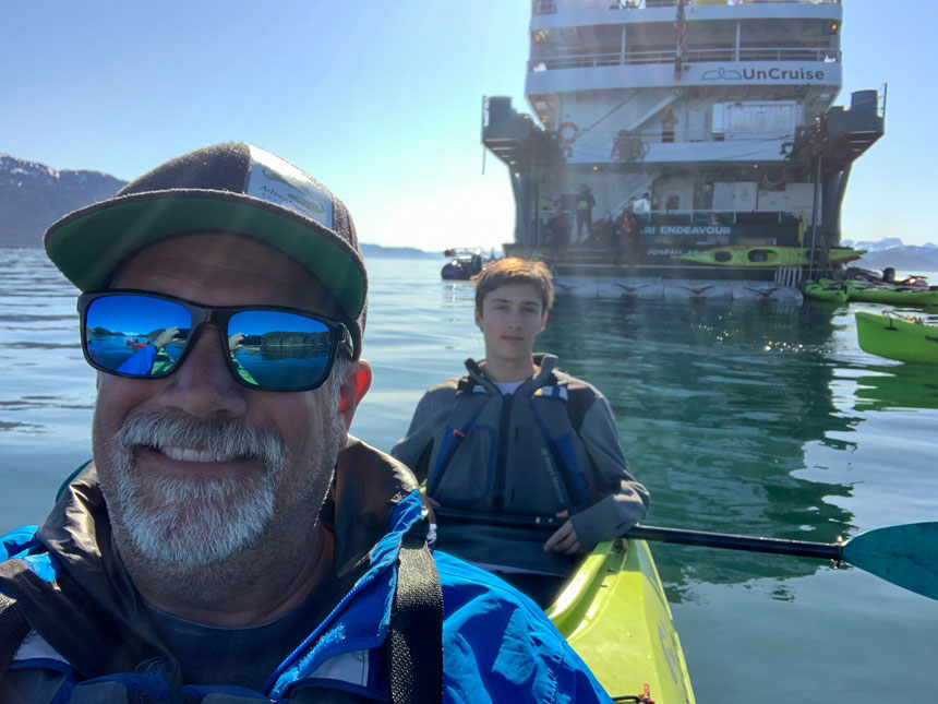 Father in sunglasses u0026 blue jacket paddles tandem kayak with son sitting behind on a sunny day in Alaska.