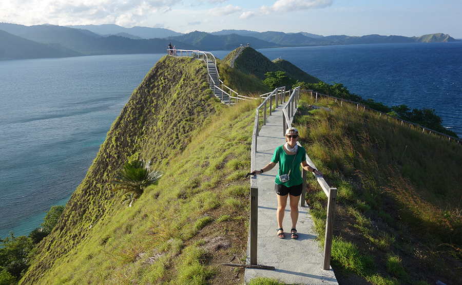 A female traveler in a green shirt standing on a wooden walkway on top of a steep grass covered hill surrounded by water.