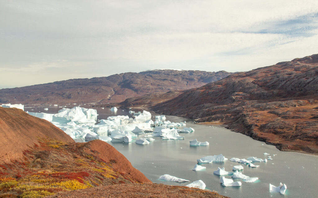 A cloudy sky blankets a view of  orange, yellow and blue toned mountains. A body of water sits between the mountains, sprinkled with icebergs in eastern Greenland. 