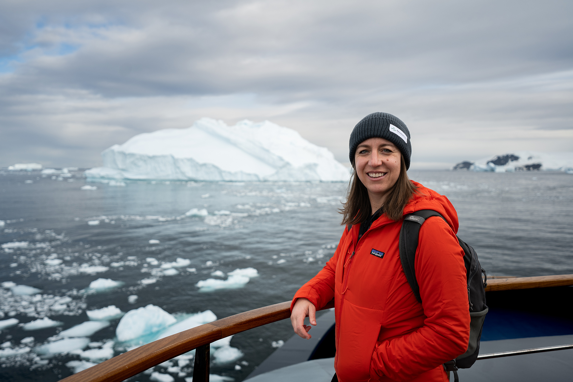Female traveler in red orange jacket and dark blue beanie leans against boat railing, massive white icebergs float in the distance.