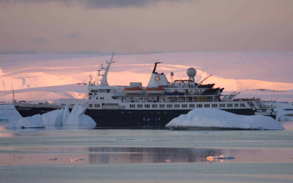A small expedition ship in Antarctica sailing through icebergs with the sun setting behind creating pink clouds.